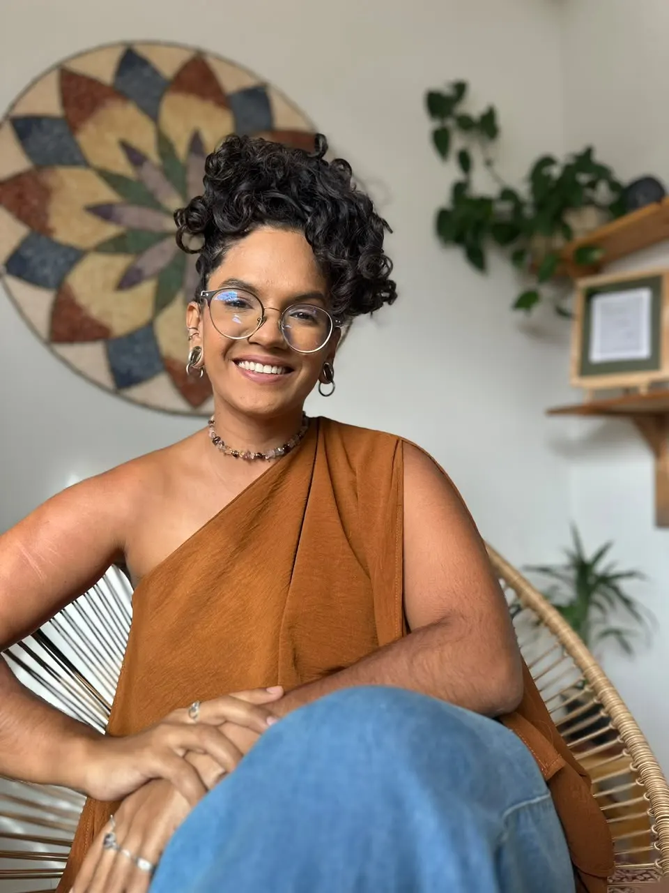 Vitória Carvalho sorrindo, sentada numa poltrona de fibra com uma mandala ao fundo